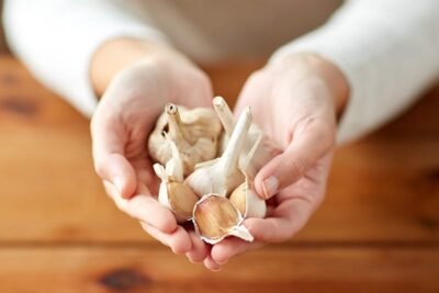 Hands holding several whole garlic bulbs and cloves over a wooden table.