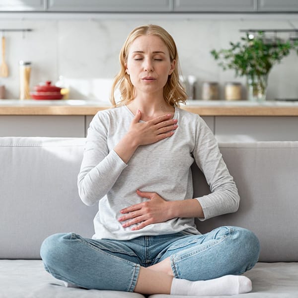 Woman sitting on couch and doing calming breathing exercises. She has a hand on her chest and another on her stomach.