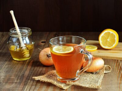 Glass of onion and lemon tea on a coaster with lemons, onion, a jar of honey in the background.