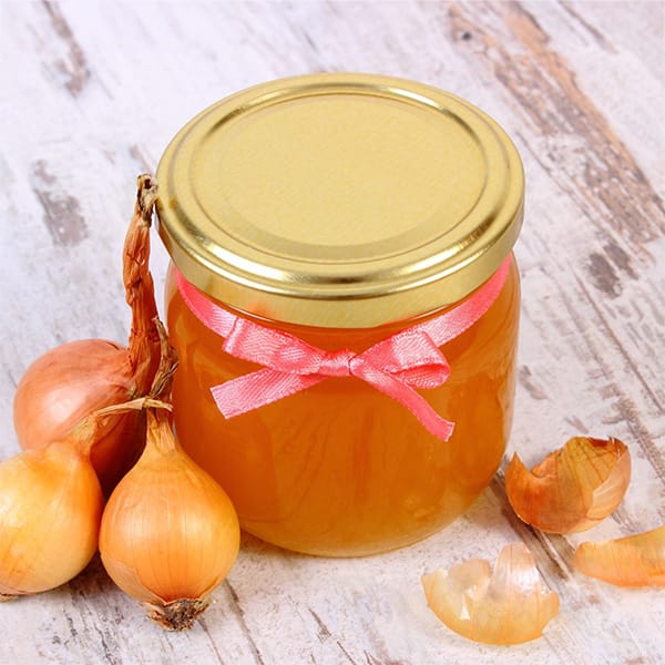 Onion cough syrup in a glass jar with a red ribbon tied around it on an old rustic wooden background. There are small onions and peels around the jar.