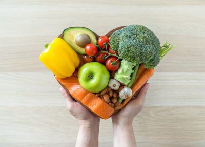 Hands holding a heart-shaped plate filled with various healthy foods like fruits and vegetables