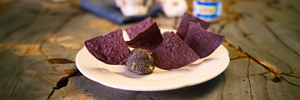 Blue corn tortilla chips served with a ball of poppy seed-coated cheese on a white plate