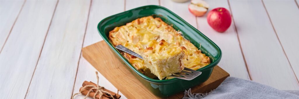 Serving of cheesy baked casserole on a wooden table with apples and cinnamon sticks nearby