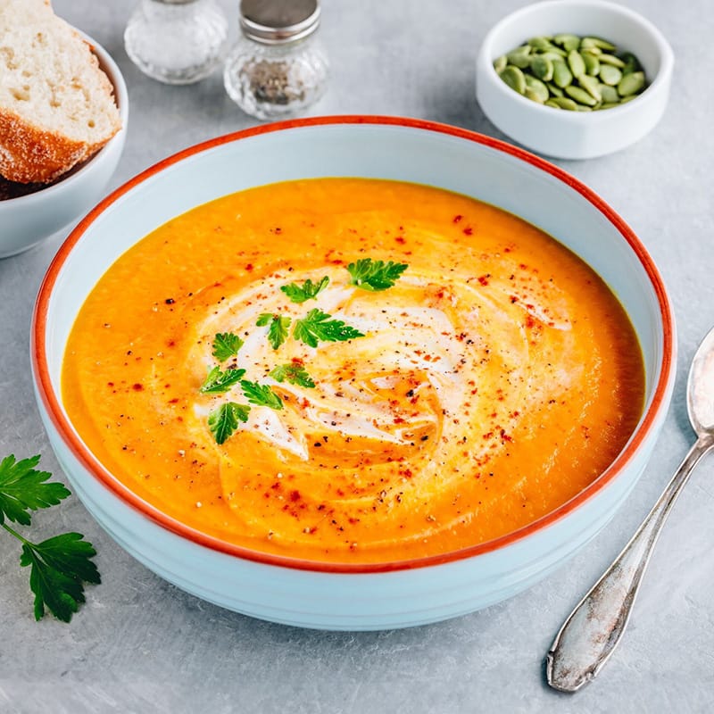 A bowl of creamy pumpkin soup garnished with herbs, served with bread and seeds on the side