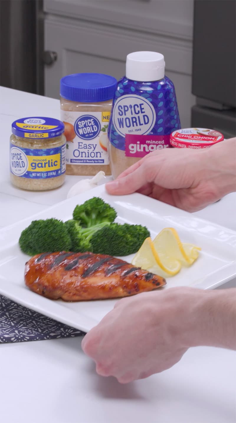 Person serving a plate of grilled chicken, broccoli, and lemon slices, with jars of spices nearby