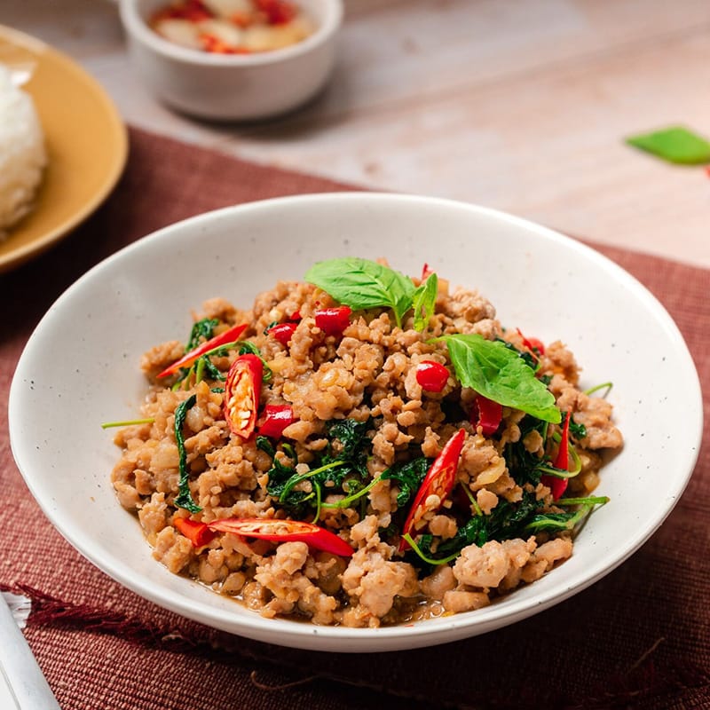 A bowl of spicy Thai basil chicken, garnished with fresh basil and chili slices
