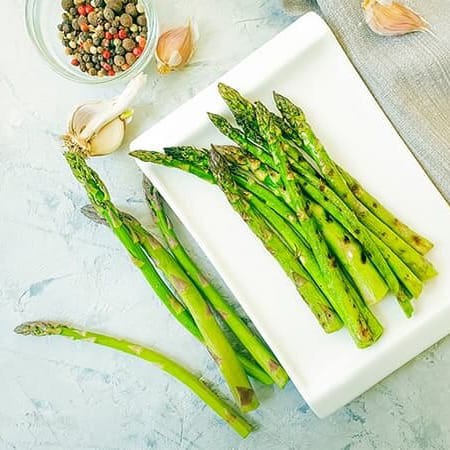 Roasted asparagus surrounded by ingredients. There are peppercorns and fresh garlic cloves.