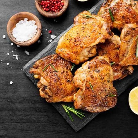Golden-brown chicken thighs topped with fresh herbs on a black cutting board surrounded by seasonings and lemon.