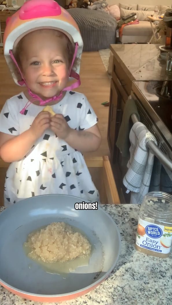 Child wearing a helmet, smiling while cooking onions in the kitchen