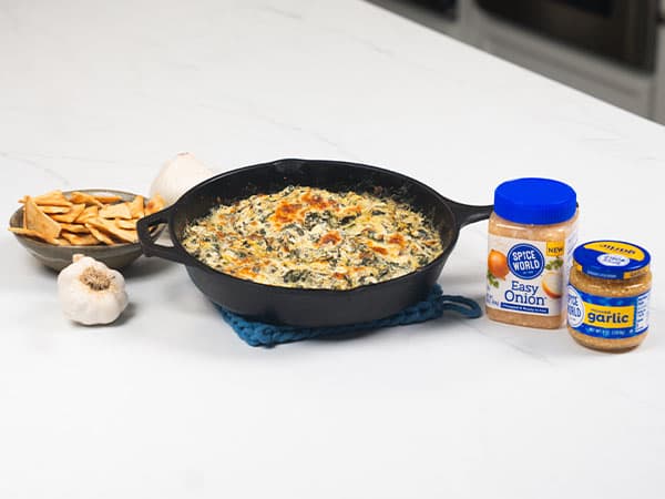 A skillet of spinach dip next to spices and pita chips on a kitchen counter