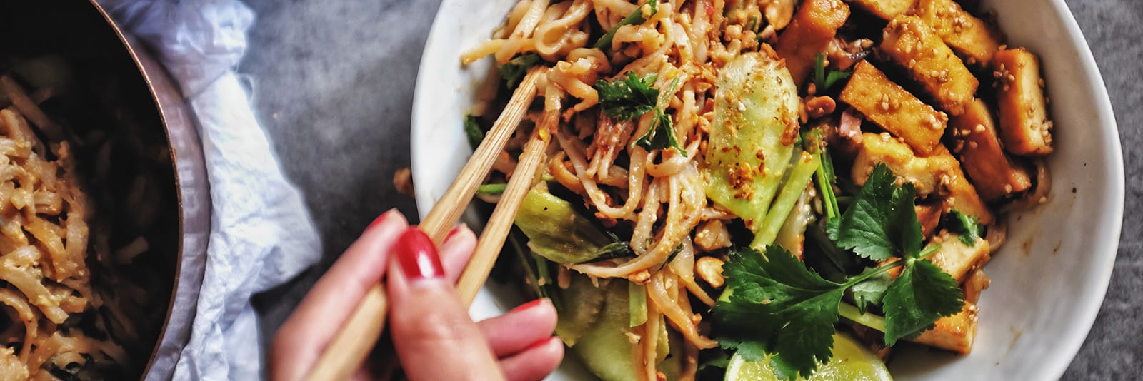 Asian stir-fry noodle bowl with tofu, bok choy, and fresh herbs being eaten with chopsticks