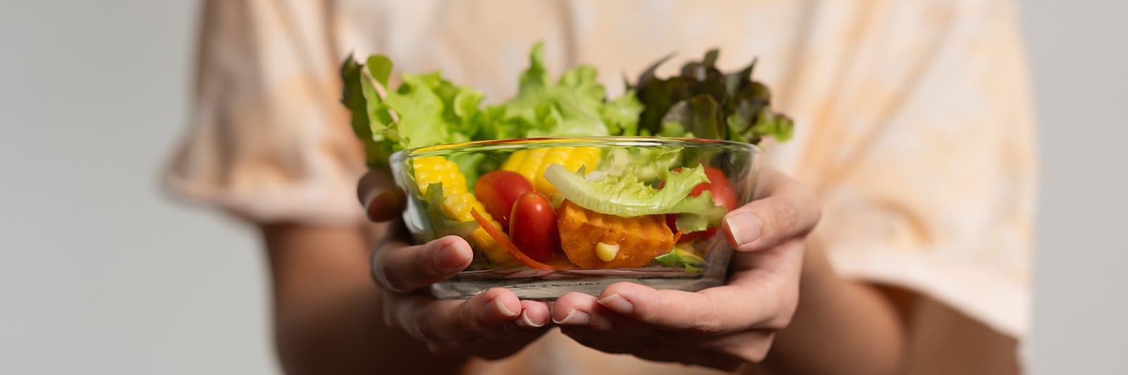 Person in beige sweater holding glass bowl of fresh colorful salad against minimal background