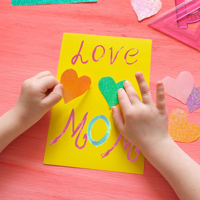 Child's hands arranging colorful heart decorations on a yellow craft paper with "love mom" written on it