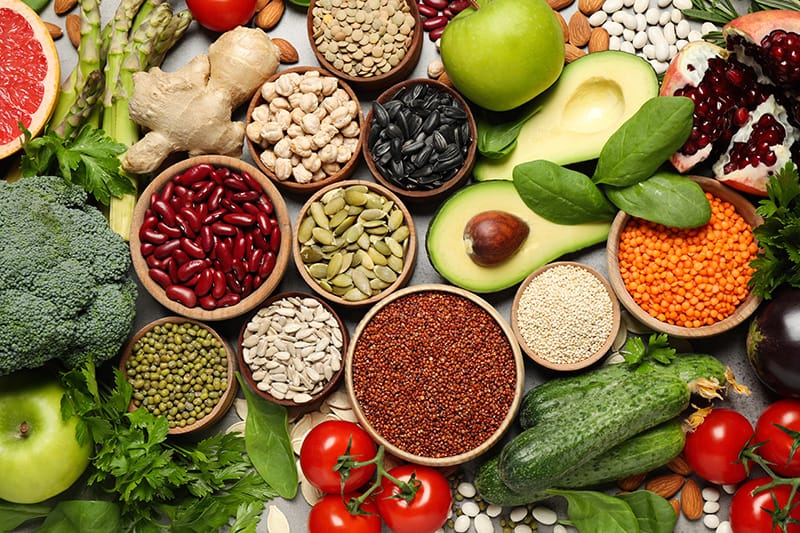 Variety of plant-based organic ingredients including legumes, nuts, seeds, avocado, and vegetables arranged in wooden bowls on gray background
