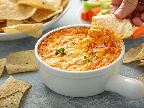 Hand dipping a chip into a bowl of creamy, cheesy dip, surrounded by vegetables and extra chips