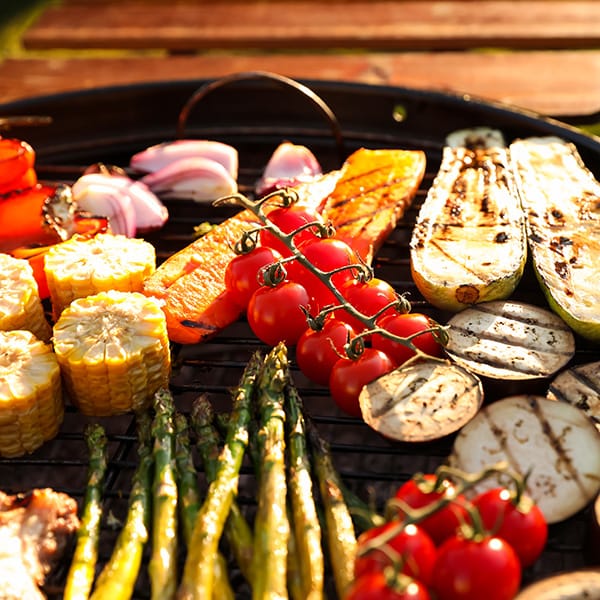 Assorted vegetables including tomatoes, corn, and asparagus grilling on a barbecue