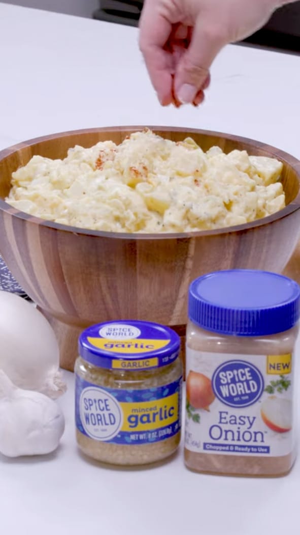 Hand seasoning a bowl of creamy potato salad, with jars of garlic and onion spices nearby