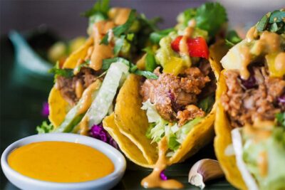 Three tacos filled with meat and vegetables, served with a side of spicy sauce