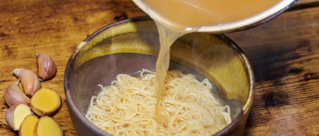 Pouring broth into a bowl of noodles, with garlic and ginger on the side