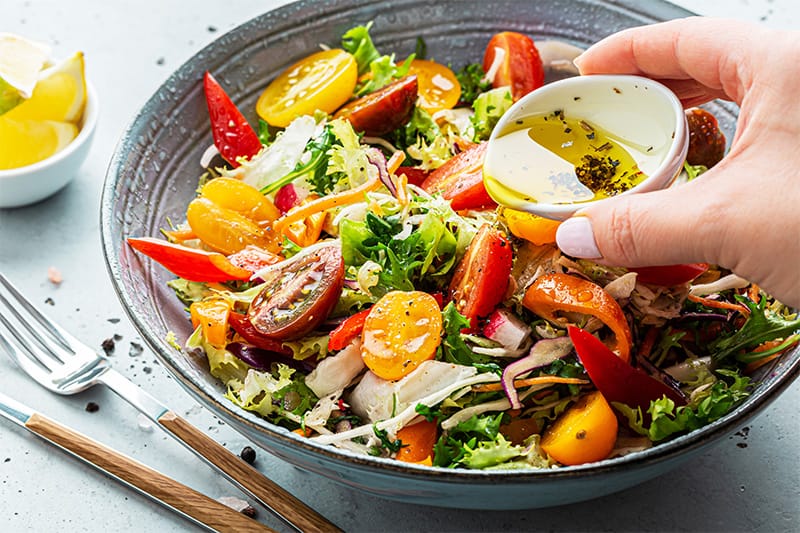 Hand drizzling homemade dressing over fresh summer salad with colorful vegetables.
