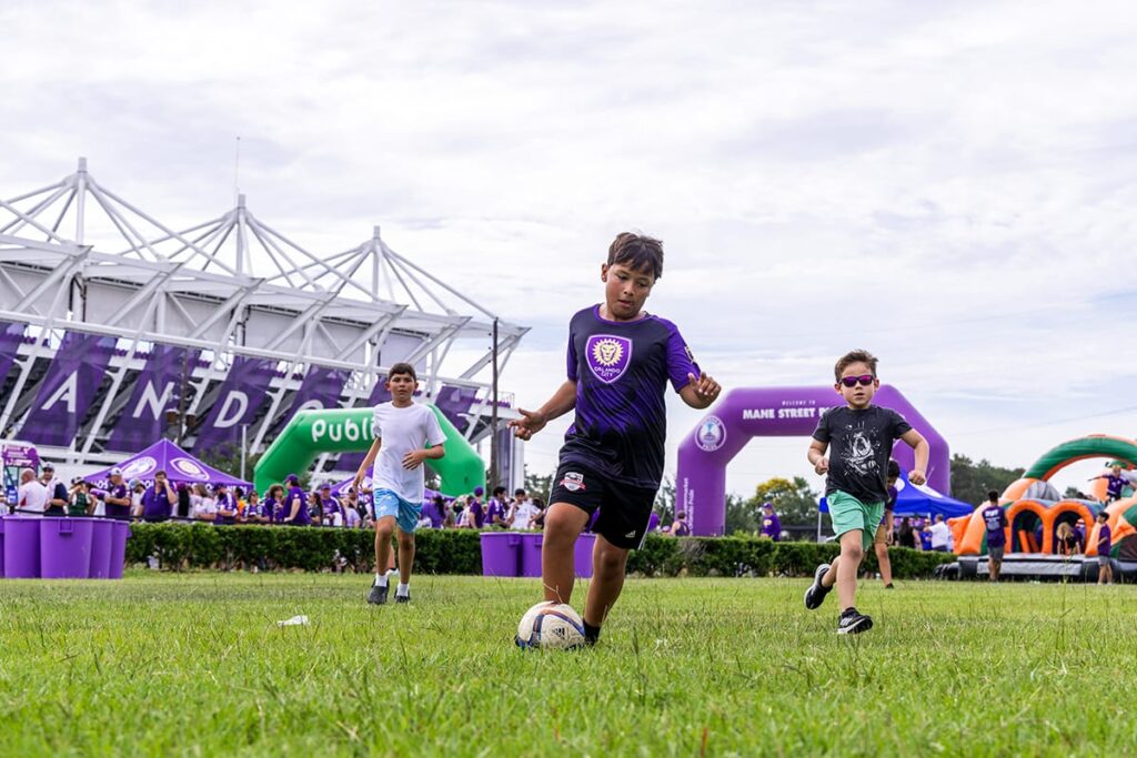 Children playing soccer in a park with event banners in the background