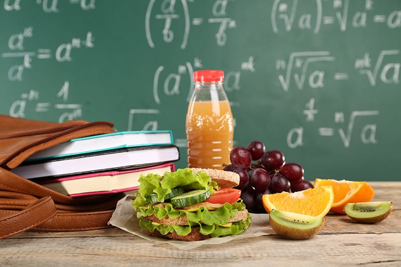Healthy school lunch with sandwich, fruits, and juice on a desk with a chalkboard background