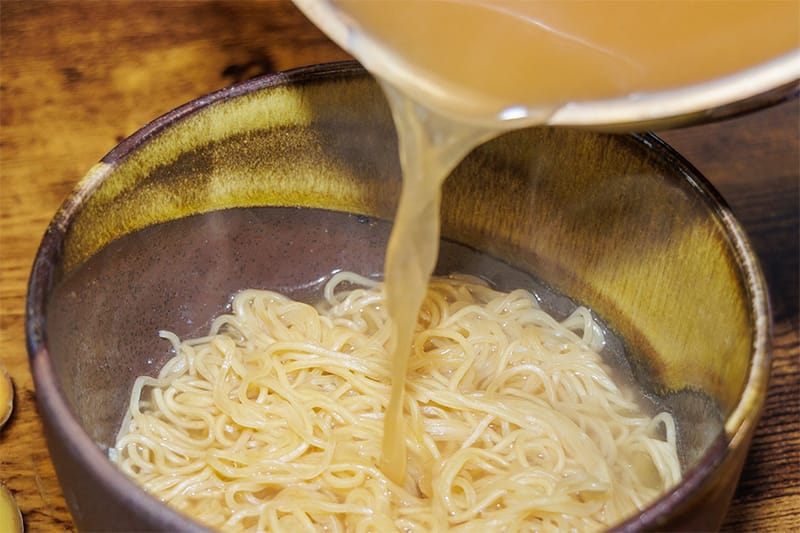 Broth being poured into a bowl of noodles on a wooden table