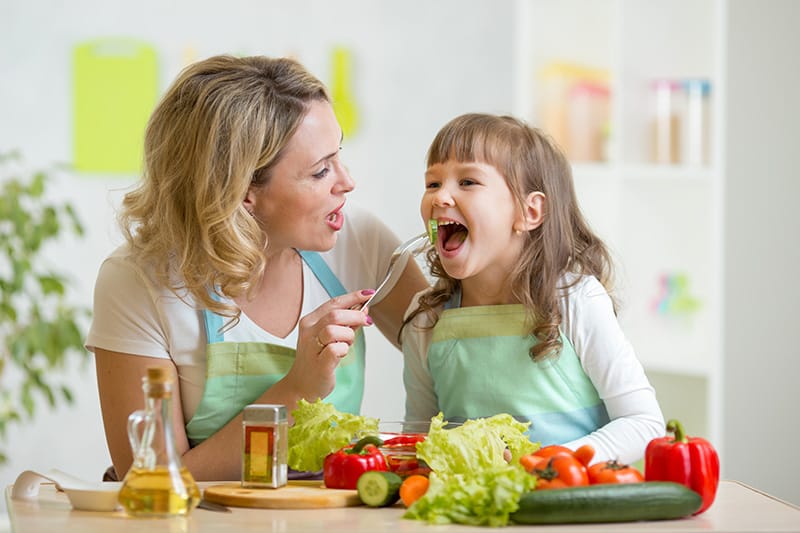 Smiling mother in an apron feeding her young daughter a piece of cucumber while preparing a meal in the kitchen.