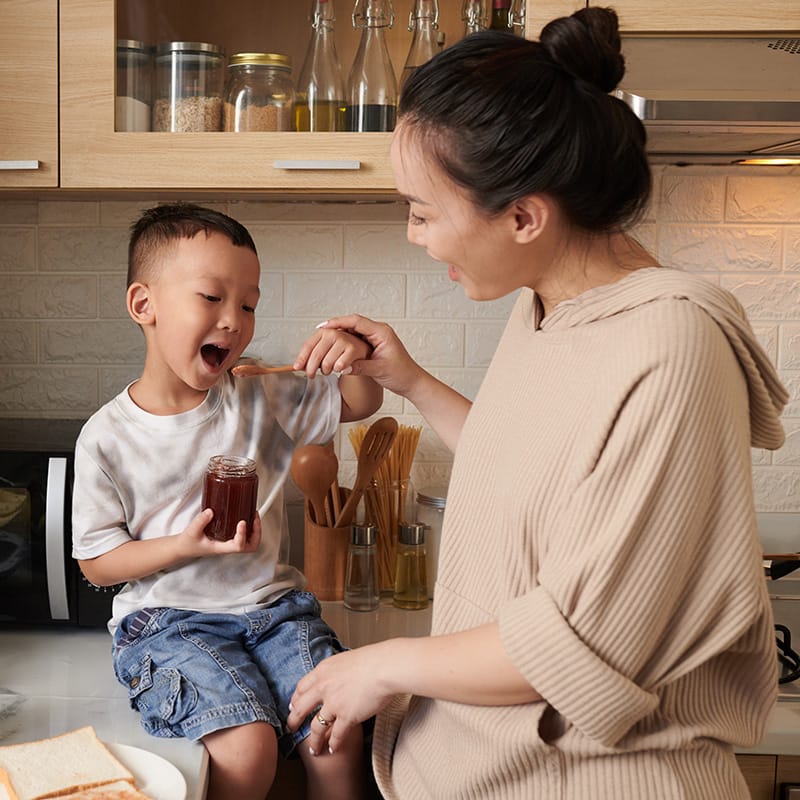 Mother feeding her son a spoonful of honey in the kitchen while he holds the jar and smiles.