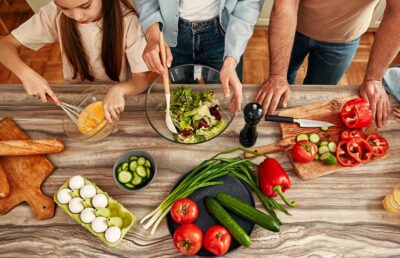 A child whisks eggs while two adults prepare a salad with cucumbers, tomatoes, peppers, and bread on the counter.