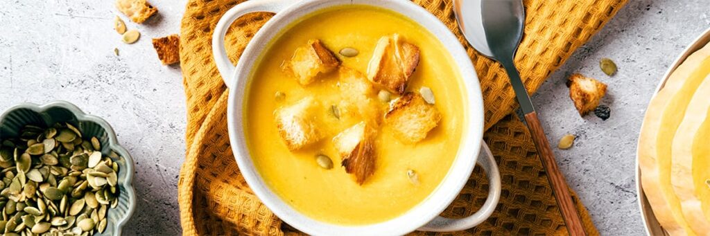 A bowl of creamy pumpkin soup garnished with croutons and pumpkin seeds, served with a spoon
