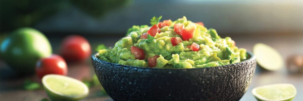 Bowl of fresh guacamole topped with tomatoes, surrounded by lime and ingredients for a tasty dip