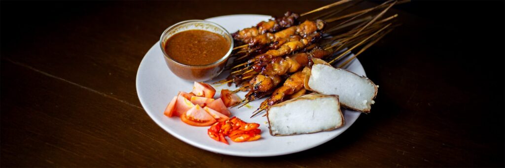 Plate of skewered meat with dipping sauce, rice cakes, and fresh vegetables, inviting a delicious dining experience