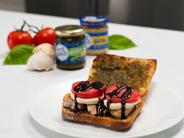 A delicious sandwich topped with chicken, tomatoes, and balsamic glaze, served with fresh ingredients in the background